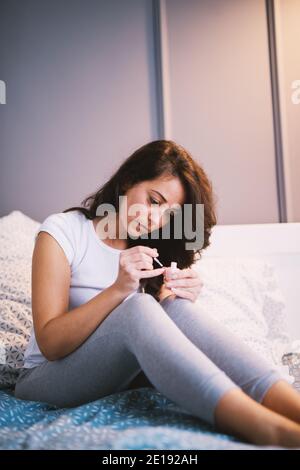 Attraktive wunderschöne Frau mittleren Alters macht Maniküre für sich selbst, während auf dem Bett im Schlafanzug sitzen. Stockfoto