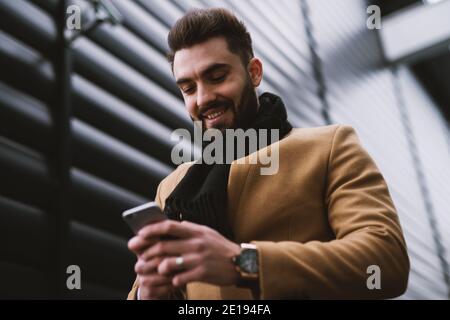 Hübscher junger Mann lächelt, während er auf dem Telefon beim Gehen auf der Straße schaut. Stockfoto