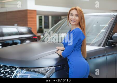 Wunderschöne glückliche rothaarige Frau, die in der Nähe ihres neuen Autos im Autohaus steht, Platz kopieren Stockfoto