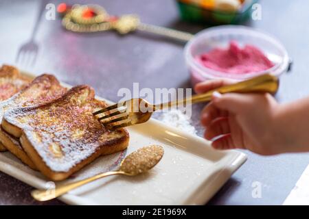 Eine kleine Hand klebt eine Gabel in den traditionellen french Toast mit Zimt auf einem grauen Hintergrund. Traditionelle spanische süß gebratene Toasts, Torrijas, mit Banane und Kaki, frische Sahne, Puderzucker auf einem Keramikplatte und Goldbesteck. Draufsicht. Hochwertige Fotos Stockfoto