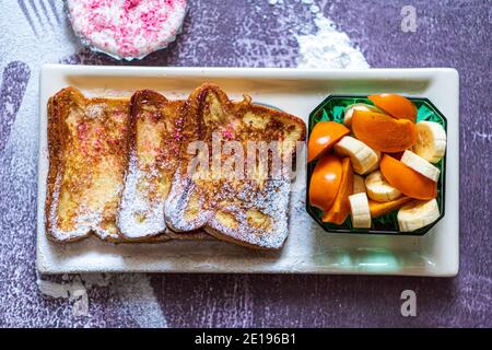 Traditioneller französischer Toast mit Zimt auf grauem Hintergrund. Traditionelle spanische süß gebratene Toasts, Torrijas, mit Banane und Kaki, frische Sahne, Puderzucker auf einem Keramikplatte und Goldbesteck. Typisches Dessert für Fastenzeit und Ostern. Draufsicht. Hochwertige Fotos Stockfoto