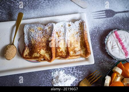 Traditioneller französischer Toast mit Zimt auf grauem Hintergrund. Traditionelle spanische süß gebratene Toasts, Torrijas, mit Banane und Kaki, frische Sahne, Puderzucker auf einem Keramikplatte und Goldbesteck. Typisches Dessert für Fastenzeit und Ostern. Draufsicht. Hochwertige Fotos Stockfoto