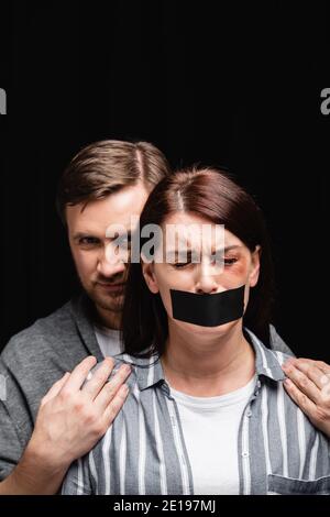 Missbräuchlicher Ehemann umarmt depressive Frau mit Klebeband und Prellung Isoliert auf Schwarz Stockfoto