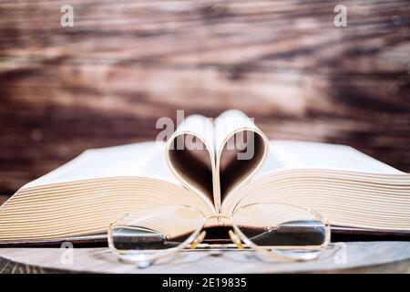 Altes geöffnetes Hardback-Buch mit Herzbogen und Lesebrille, isoliert auf einem Holzhintergrund. Ich lese gerne. Nahaufnahme Herz aus Buch Stockfoto