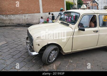 Antananarivo, Madagaskar - 24. April 2019: Weißes Taxiauto, das langsam über Fliesensteinstraße fährt, Gruppe unbekannter madagassischer Menschen im Hintergrund. Madagas Stockfoto