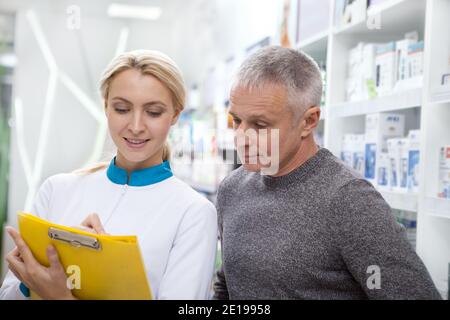 Attraktive junge Apothekerin hilft reifen Mann einkaufen für Medikamente in der Apotheke. Schöne weibliche Chemikerin, die in der Apotheke arbeitet und gibt Stockfoto
