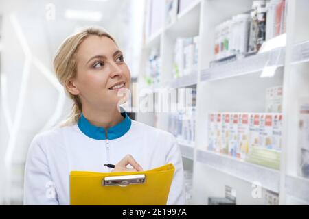 Attraktive, fröhliche Apothekerin, die den Bestand in einem Gang überprüft und Notizen auf ihr Klemmbrett macht. Schöner Apotheker lächelt freudig, arbeitet an der Stockfoto