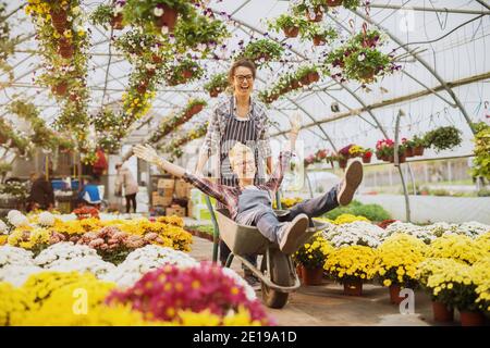 Zwei fröhlich lächelnde Floristen Frauen Arbeiter genießen eine Pause und fahren in einen Wagen im Gewächshaus voller bunten Blumen und Töpfe. Stockfoto