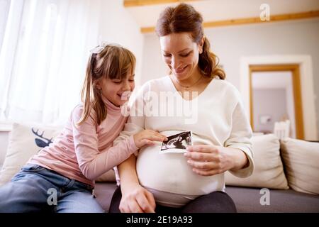 Süße fröhliche kleine Kleinkind Mädchen sitzt auf dem Sofa und Blick auf Ultraschall-Scan-Bild, während ihre schöne schwangere Mutter Halten Sie den Bauch fest Stockfoto