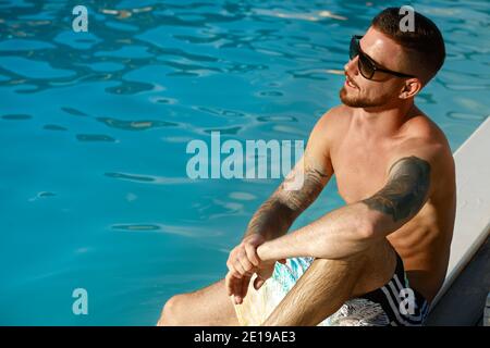 Schöner tätowierte bärtiger Mann, der an einem heißen, sonnigen Tag am Pool relaxt. Attraktiver junger Mann, der seinen Sommerurlaub genießt, sich am Pool ausruhend, Cop Stockfoto