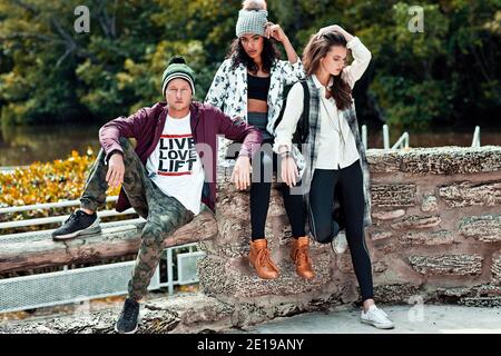 Porträt von 3 jungen Erwachsenen, die an einem sonnigen Frühlingstag vor einem See in einem Naturpark sitzen und coole urbane Outfits tragen. Stockfoto