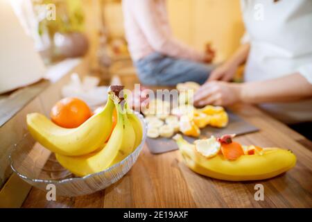 Nahaufnahme Fokus Ansicht von Bananen und Orangen während der Schwangerschaft Frau bereitet Obstsalat, während ihre hübsche Kleinkind Tochter sitzen Auf dem Küchentisch n Stockfoto
