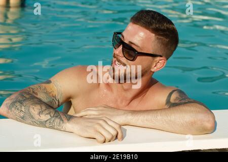 Nahaufnahme eines hübschen bärtigen Mannes in Sonnenbrillen lächelnd wegschauen, während Sie sich am Pool entspannen. Glücklicher Mann, der sich nach dem Schwimmen ausruhte. Stockfoto