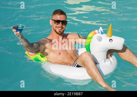 Fröhlicher, schöner bärtiger Mann, der mit seinem Cocktailglas jubelt, während er am Pool auf einem aufblasbaren Einhorn schwimmt. Glücklicher Mann, der sich während der Hallo erholte Stockfoto