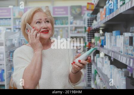 Charmante ältere Frau lächelt freudig und spricht am Telefon, während sie Kosmetikprodukte in der Drogerie kauft. Fröhliche ältere Frau mit ihrem intelligenten p Stockfoto