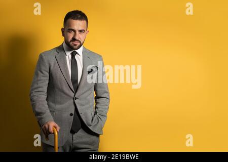 Old School trendy stilvolle schöne junge Mann trägt grauen Anzug Mit einer Hand angehoben stehend mit Stock oder Schirm in Seine Hand isoliert auf gelb Stockfoto