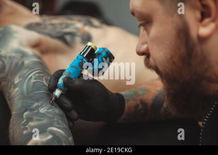 Nahaufnahme eines bärtigen professionellen Tätowierers, der konzentriert aussieht, während er einen jungen Mann auf seinem Arm tätowiert. Talentierter Tätowierer, der daran arbeitet Stockfoto