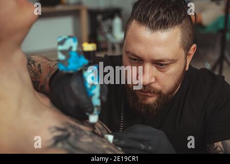 Professionelle Tattoo-Künstler suchen konzentriert, arbeiten in seinem Studio, machen ein Tattoo auf der Brust seines Kunden. Bärtiger Tätowierer, der ein Tattoo für h macht Stockfoto