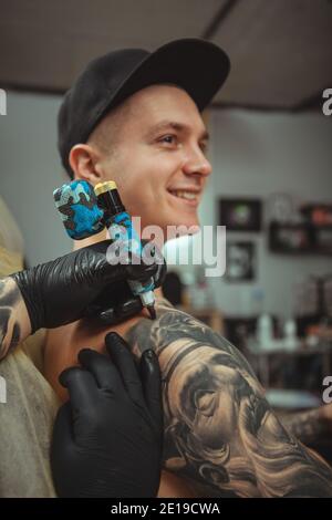 Vertikale Nahaufnahme eines hübschen jungen Mannes, der lächelt und auf seinen Arm tätowiert wird. Zugeschnittenes Schuss von Händen von professionellen Tattoo-Künstler arbeiten, machen ein Stockfoto