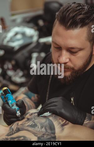 Vertikale Aufnahme eines bärtigen professionellen Tatoo-Künstlers, der sich konzentriert, während er in seinem Studio arbeitet. Zugeschnittene Aufnahme eines Tätowierers Zeichnung Tattoo auf Brust o Stockfoto