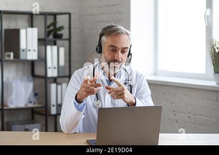 Porträt eines älteren grauhaarigen männlichen Arztes in seinem Büro mit Laptop für Video-Chat mit einem Patienten. Online-Beratung mit Arzt für Diagnosen und Stockfoto