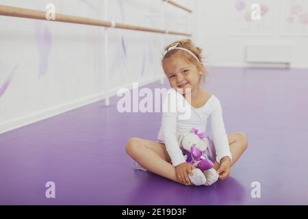 Entzückende kleine Ballerina lächelt fröhlich zur Kamera, sitzt auf dem Boden im Tanzstudio mit ihrem Spielzeug. Nettes kleines Mädchen Ruhe nach dem Tanzen cla Stockfoto