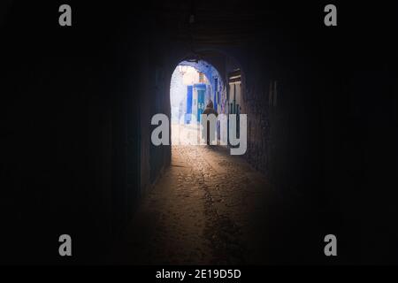 Ein Mann mit einer traditionellen marokkanischen Djellaba geht durch einen dunklen Tunnel und Torbogen in Chefchaouen, Marokko, ins Licht. Stockfoto