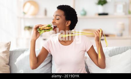 Afroamerikanische Frau, die Cheeseburger essen will, zieht sich zu Hause mit Maßband weg, Panorama Stockfoto