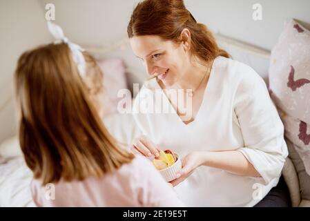 Rückansicht von kleinen Kleinkind Mädchen bringt einen Obstsalat in einer weißen Tasse zu ihrer überrascht schwangere Mutter. Familie und neues Lebenskonzept. Stockfoto