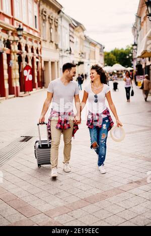 Schöner Mann mit Reisetasche hält Hände mit niedlichen Mädchen mit Hut beim Spaziergang durch die Stadt. Stockfoto