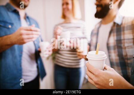 Gruppe von jungen Geschäftsleuten, die miteinander reden und Pappbecher Kaffee halten. Fokusansicht von Hand und Papiertasse. Stockfoto