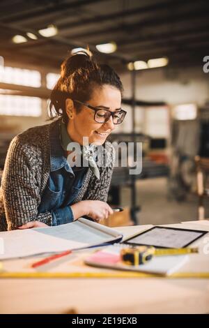 Nahaufnahme der charmanten lächelnden motivierten Ingenieurin mittleren Alters mit Brillen, die in der Werkstatt mit Blaupausen und Tablet arbeiten. Stockfoto