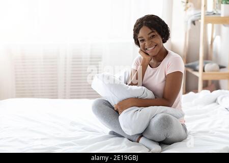 Schöne schwarze Frau im Schlafanzug sitzt auf dem Bett, umarmende Kissen Stockfoto