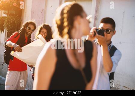 Junger Mann, der mit einer professionellen Kamera eine Freundin fotografiert, während zwei andere im Hintergrund nach einer Richtung auf einer Karte suchen. Stockfoto