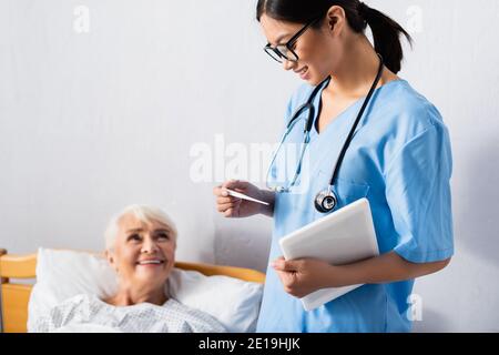 Lächelnde asiatische Krankenschwester mit digitalen Tablet Blick auf Thermometer in der Nähe Glückliche ältere Frau auf verschwommenem Hintergrund Stockfoto