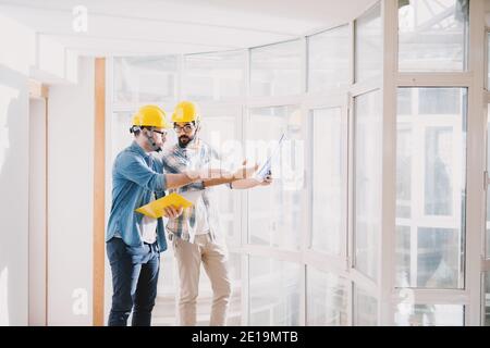 Zwei fokussierte junge professionelle Ingenieure mit Helmen, die im schönen großen hellen Büro mit großen Fenstern eine Diskussion über das Projekt führen. Stockfoto