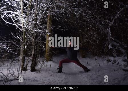 Yoga üben im Winter, im Wald bei Nacht im Schnee. Neue Herausforderungen mit Schnee und Kälte bringen dir bei, wie du Fokus und Kontrolle erreichst. Stockfoto