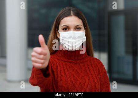 COVID-19 optimistische junge Frau mit Schutzmaske zeigt Daumen nach oben In der Stadt Straße bereit für die Impfkampagne gegen Coronavirus Diseas Stockfoto