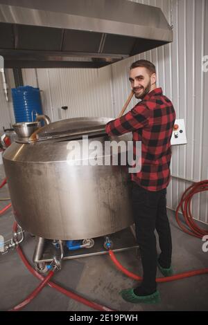 Fröhlicher Brauer lächelt zur Kamera, während er die Biergärung kontrolliert Prozess in seiner Mikrobrauerei Stockfoto