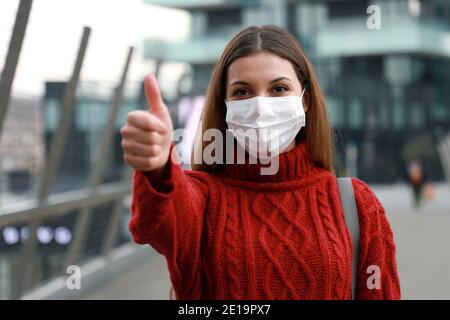 Optimistische junge Frau trägt Schutzmaske zeigt Daumen nach oben in City Street bereit für die Impfkampagne gegen Coronavirus-Krankheit 2019 Stockfoto