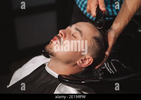 Kurzer Schuss eines reifen Mannes, der seine Haare vom professionellen Friseur waschen ließ. Nicht erkennbarer Barbier, der im Friseurladen arbeitet und Haare eines Kunden wäscht Stockfoto