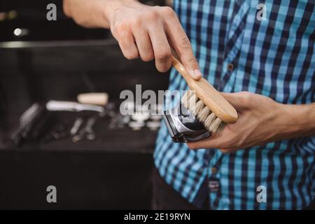 Ausschnitt aus der Nähe eines professionellen Barbiers, der seinen elektrischen Haarschneider nach der Arbeit mit einer Bürste reinigt, kopieren Sie den Platz. Friseur kümmert sich um seinen Hairsty Stockfoto