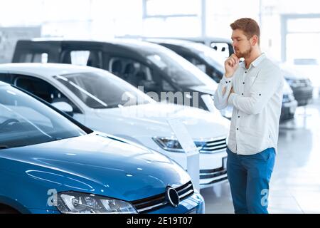 Endgültige Entscheidung. Aufnahme eines hübschen jungen Mannes, der vor einem neuen Auto beim Autohaus steht und denkt, sich das Kinn zu reiben Stockfoto