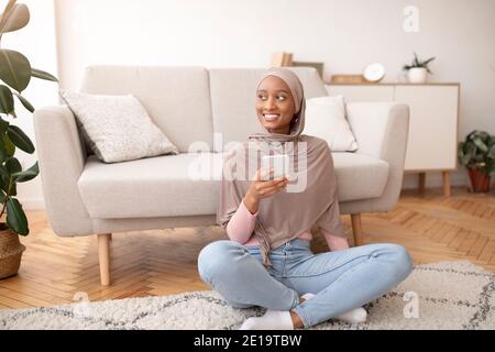 Muslimische Dame in Hijab mit Handy auf dem Boden im Wohnzimmer, Textnachrichten lesen, Surfen im Internet, im Gespräch mit einem Freund Stockfoto