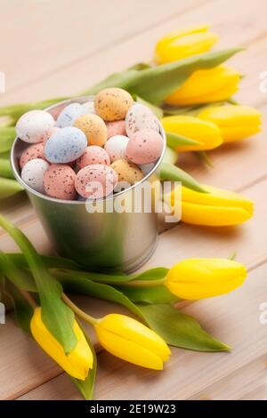 Bunte Ostereier in einem Stahleimer unter Haufen Von gelben Tulpen auf Holztisch Stockfoto