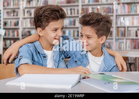 Fröhliche junge Zwillingsbrüder, die lachen und sich an der Bibliothek umarmen, während sie gemeinsam Schulaufgaben machen Stockfoto
