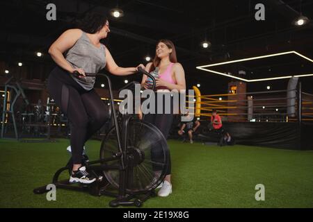 Body positive, Weiblichkeit Konzept. Übergewichtige glückliche Frau im Gespräch mit ihrem Freund beim Radfahren auf dem Air Bike im Fitnessstudio. Fröhlich plus Größe Sportwomen c Stockfoto