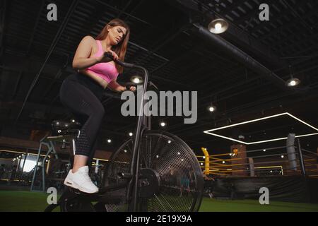 Low-Angle-Aufnahme einer Plus-Size-Frau in Sportbekleidung Radfahren auf dem Air Bike. Junge übergewichtige Sportlerin, die Cardio-Training auf dem Luftfahrrad im Fitnessstudio, CO Stockfoto