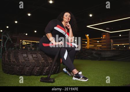 Low-Angle-Aufnahme einer Plus-Size-Frau in Sportkleidung, die nach dem Training mit Vorschlaghammer im Fitnessstudio ruht. Fröhliche übergewichtige Sportlerin entspannend afte Stockfoto