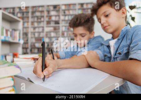 Zwillingsbrüder machen gemeinsam Hausaufgaben. Junge Jungen, die in der Bibliothek studieren, schreiben in Lehrbüchern, kopieren Raum Stockfoto
